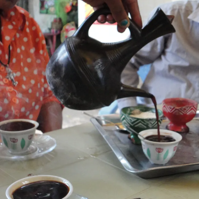 Coffee poured from a black clay jebena into small cini cups, with incense burning beside them