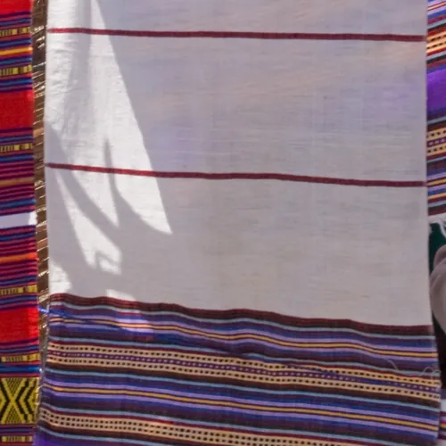 A white handwoven cotton shawl with thin red stripes and a violet woven border, hanging on a line